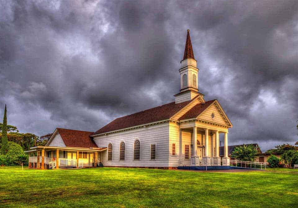 The Church at Koloa, a white historic-style church with tall columns and a steeple, illuminated beneath dramatic dark storm clouds.